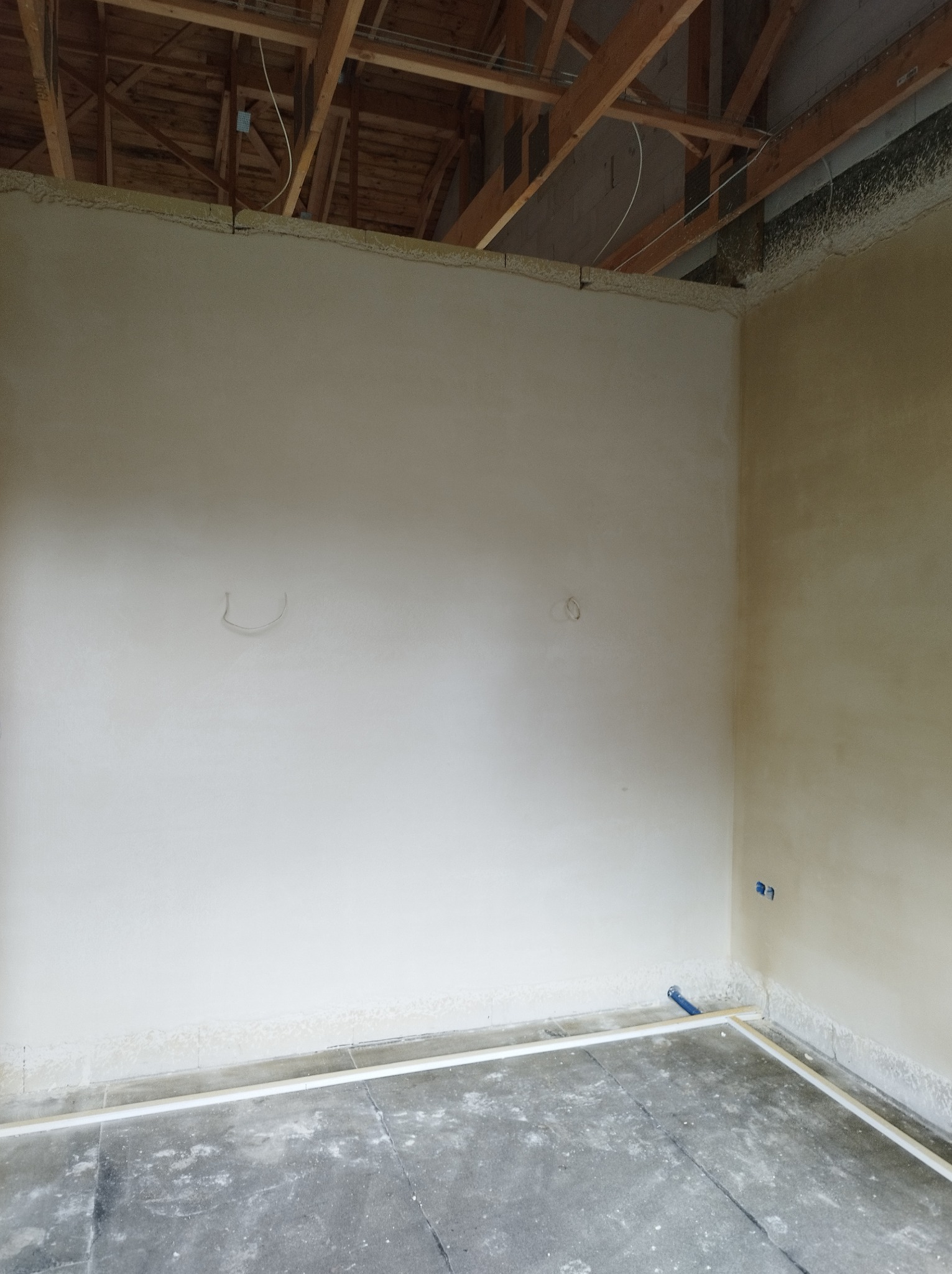 Surowe wnętrze w trakcie prac wykończeniowych: ściany pokryte wstępną warstwą tynku, widoczne ślady po instalacjach elektrycznych, sufit z odsłoniętą konstrukcją drewnianą, podłoga zabezpieczona...