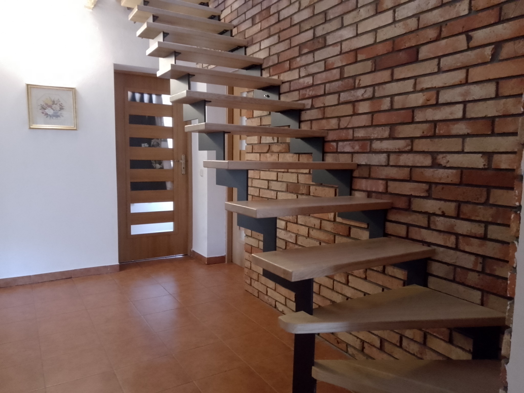 Samonośne stopnie z jasnego drewna mocowane do ściany z cegły, minimalistyczny design w nowoczesnym wnętrzu z brązowymi płytkami podłogowymi i obrazkiem na ścianie.