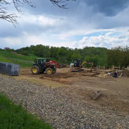 ELKAT - Przygotowanie terenu pod budowę: traktor z łyżką i mała koparka pracują na placu budowy, widoczne drewniane szalunki, paleta z kostką brukową, ubijarka i niwelator laserowy.