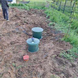 ELKAT - Dwa zielone zbiorniki na gruncie z widocznymi śladami opon, obok czerwona pokrywa, w tle ogrodzenie panelowe i osoba oddalająca się.