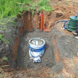 ELKAT - Wykopany dół w ziemi z zainstalowanym zbiornikiem z niebieskimi przewodami, widoczna pomarańczowa rura i żółta koparka w tle, teren budowy.