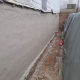 Świeżo otynkowana ściana fundamentowa z widocznym poziomem gruntu obok budynku, zabezpieczona folią i z czerwoną puszką farby w pobliżu.