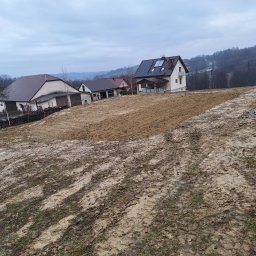 ELKAT - Przygotowany teren pod budowę z widocznymi śladami opon, otoczony domami w krajobrazie pagórkowatym, pochmurne niebo.