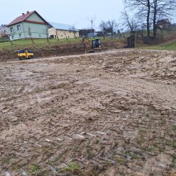 ELKAT - Przygotowanie terenu pod budowę: walec i koparka pracują na rozległym, błotnistym placu w pochmurny dzień. Widoczne ślady opon i gąsienic w grząskim gruncie.