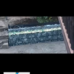 Gabionowa konstrukcja z kamienia, prawdopodobnie mur oporowy, z oświetleniem LED wzdłuż g&oacute;rnej krawędzi, częściowo zasłonięta przez roślinność i ziemię.