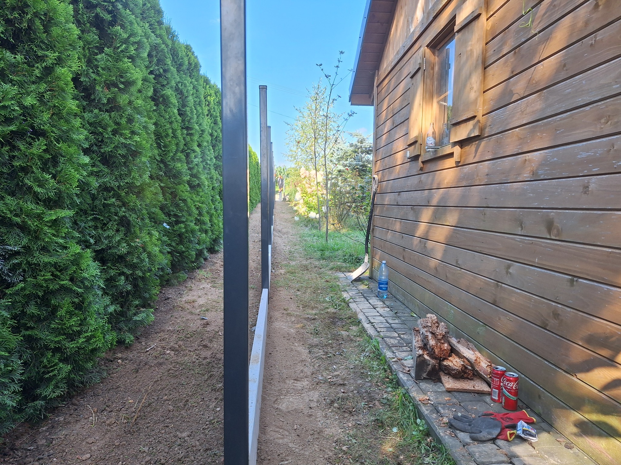 Montaż ogrodzenia: widok wzdłuż słupków i betonowej podmurówki, z tujami po lewej i drewnianym budynkiem gospodarczym po prawej stronie. Na ziemi leżą śmieci i narzędzia.