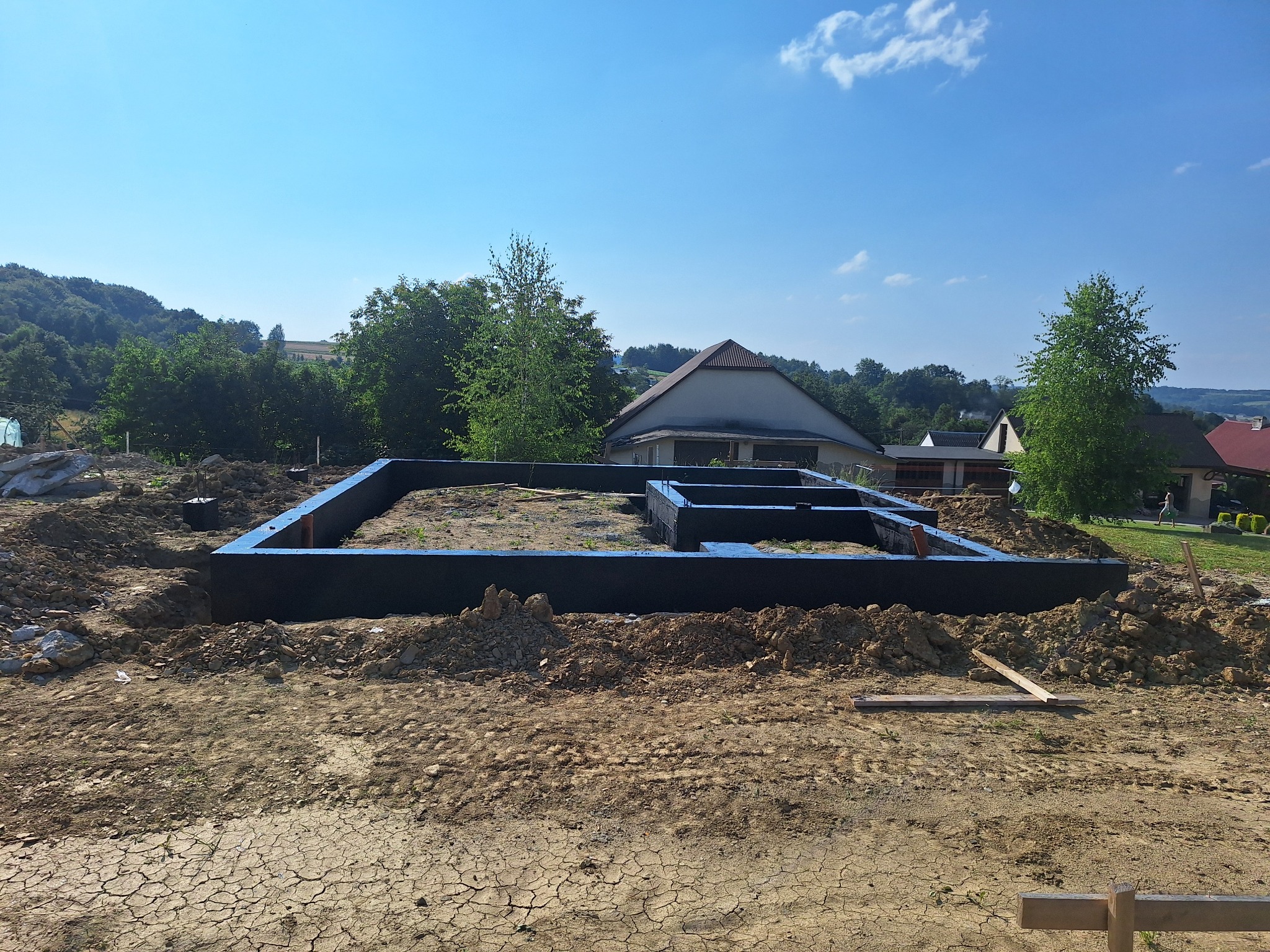 Zalane, czarne szalunki pod budowę domu, widoczne na tle wykopu z hałdami ziemi i zabudowaniami w oddali.