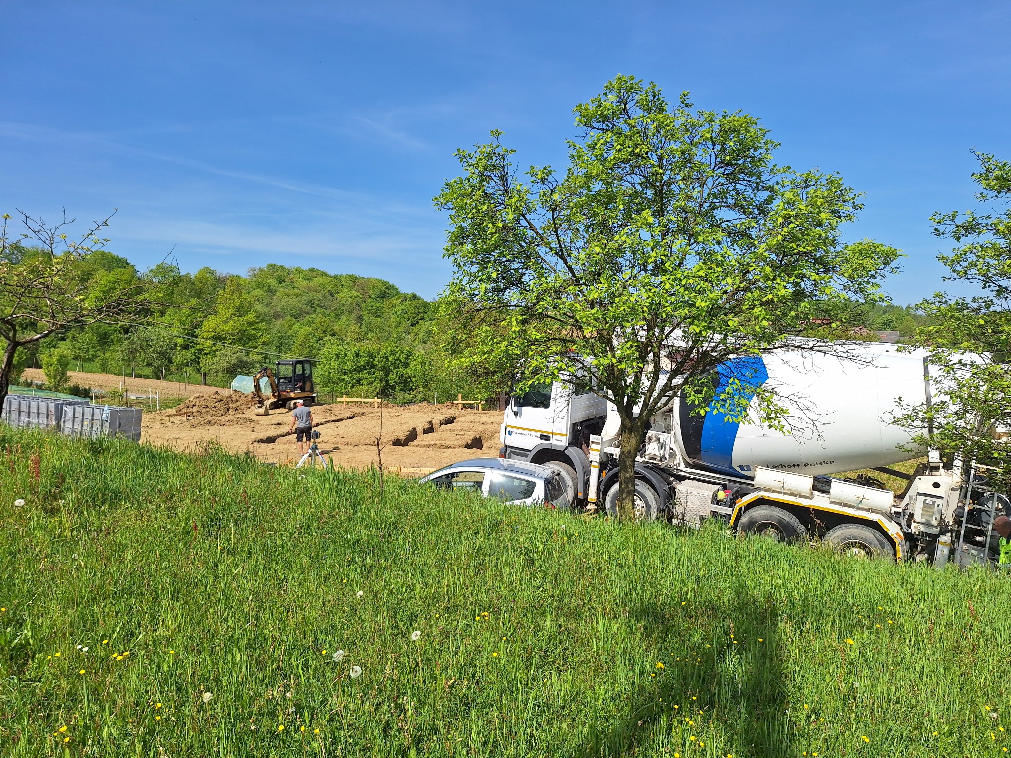 Betoniarka na tle wykopu pod fundamenty, widoczny pracownik z niwelatorem oraz koparka, teren porośnięty trawą.