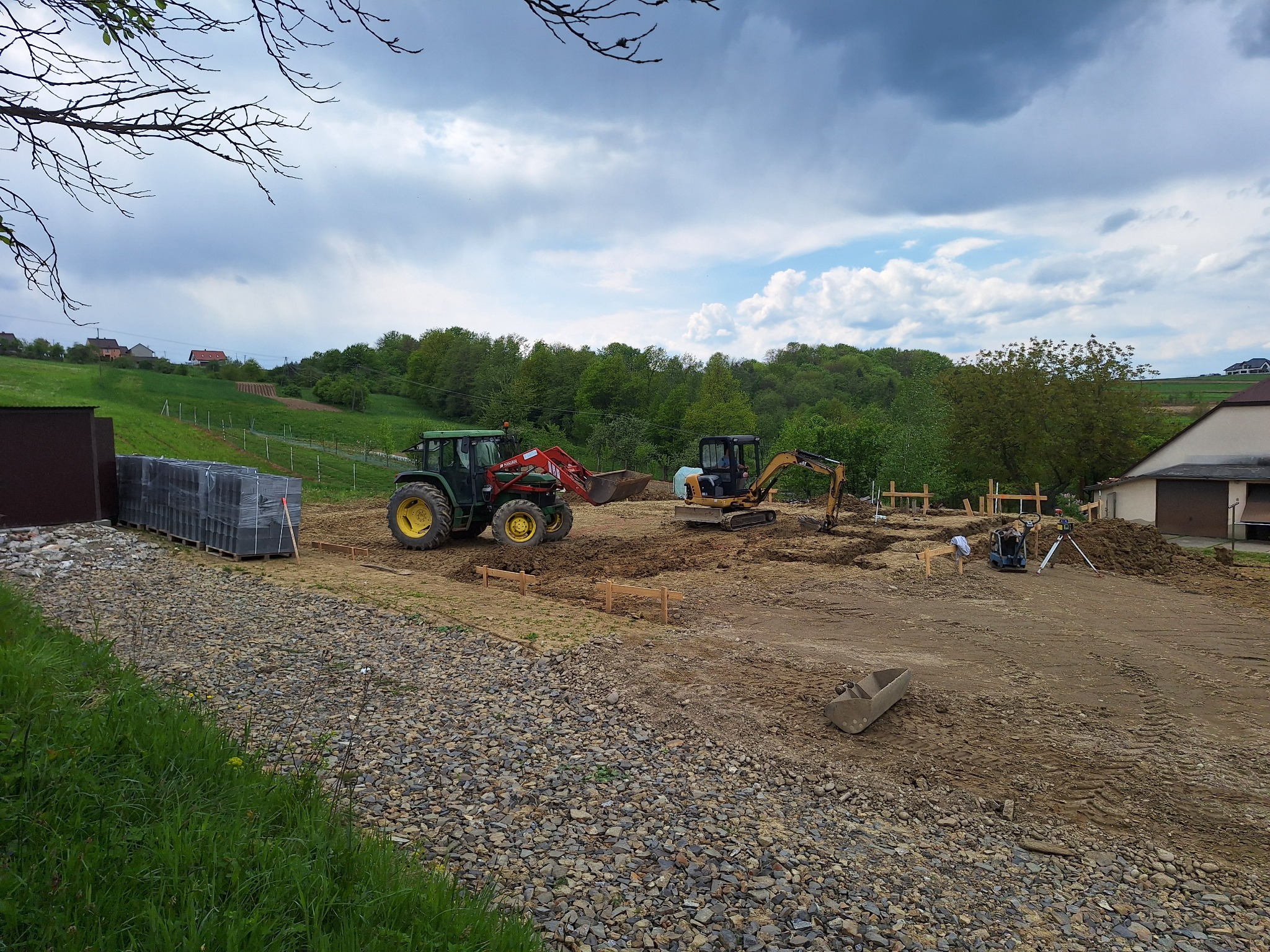 Przygotowanie terenu pod budowę: traktor z łyżką i mała koparka pracują na placu budowy, widoczne drewniane szalunki, paleta z kostką brukową, ubijarka i niwelator laserowy.