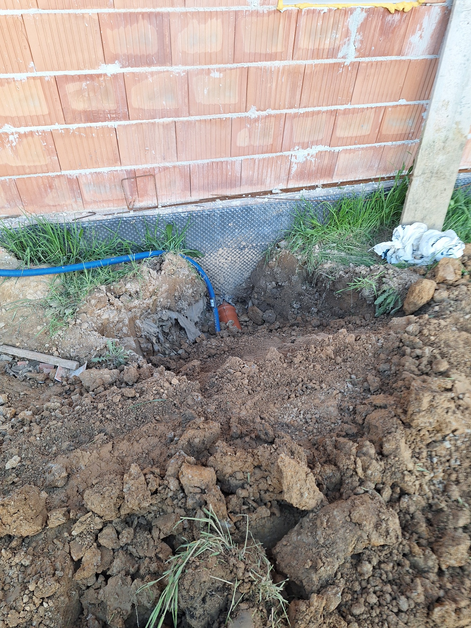 Wykop pod fundament z widoczną izolacją pionową, wystającą niebieską rurą karbowaną i fragmentem murowanej ściany z cegły ceramicznej.