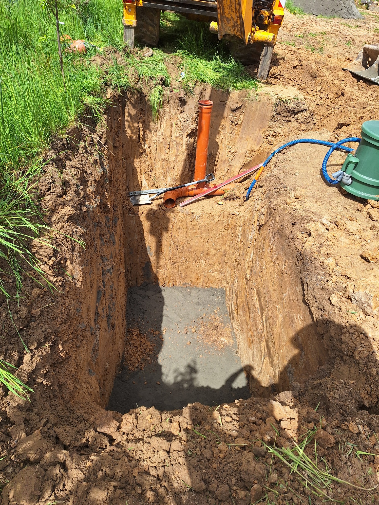 Głęboki wykop w ziemi z betonową podstawą, widoczne rury kanalizacyjne, niebieski wąż i zielony zbiornik na tle fragmentu koparki.