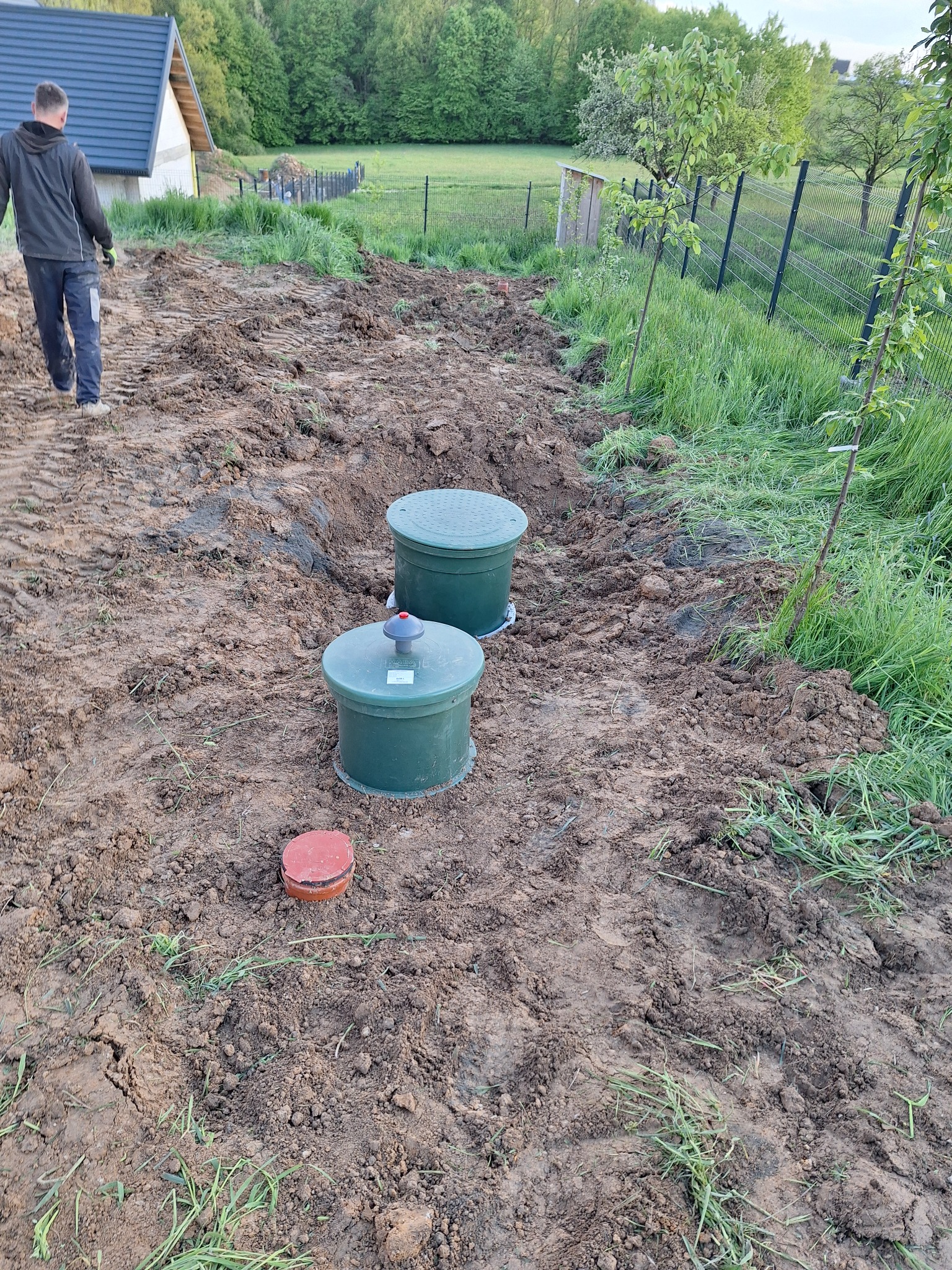 Dwa zielone zbiorniki na gruncie z widocznymi śladami opon, obok czerwona pokrywa, w tle ogrodzenie panelowe i osoba oddalająca się.