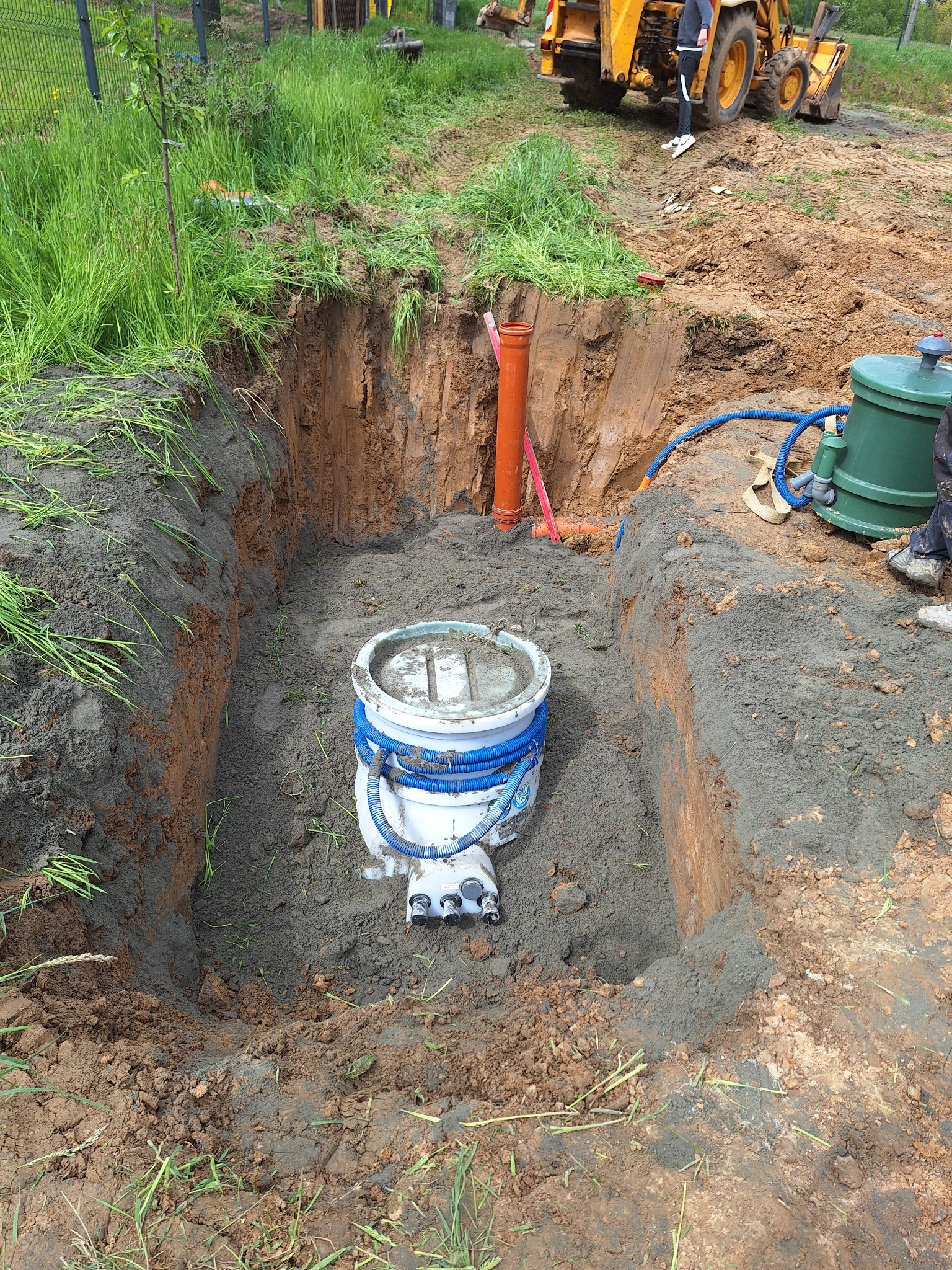 Wykopany dół w ziemi z zainstalowanym zbiornikiem z niebieskimi przewodami, widoczna pomarańczowa rura i żółta koparka w tle, teren budowy.