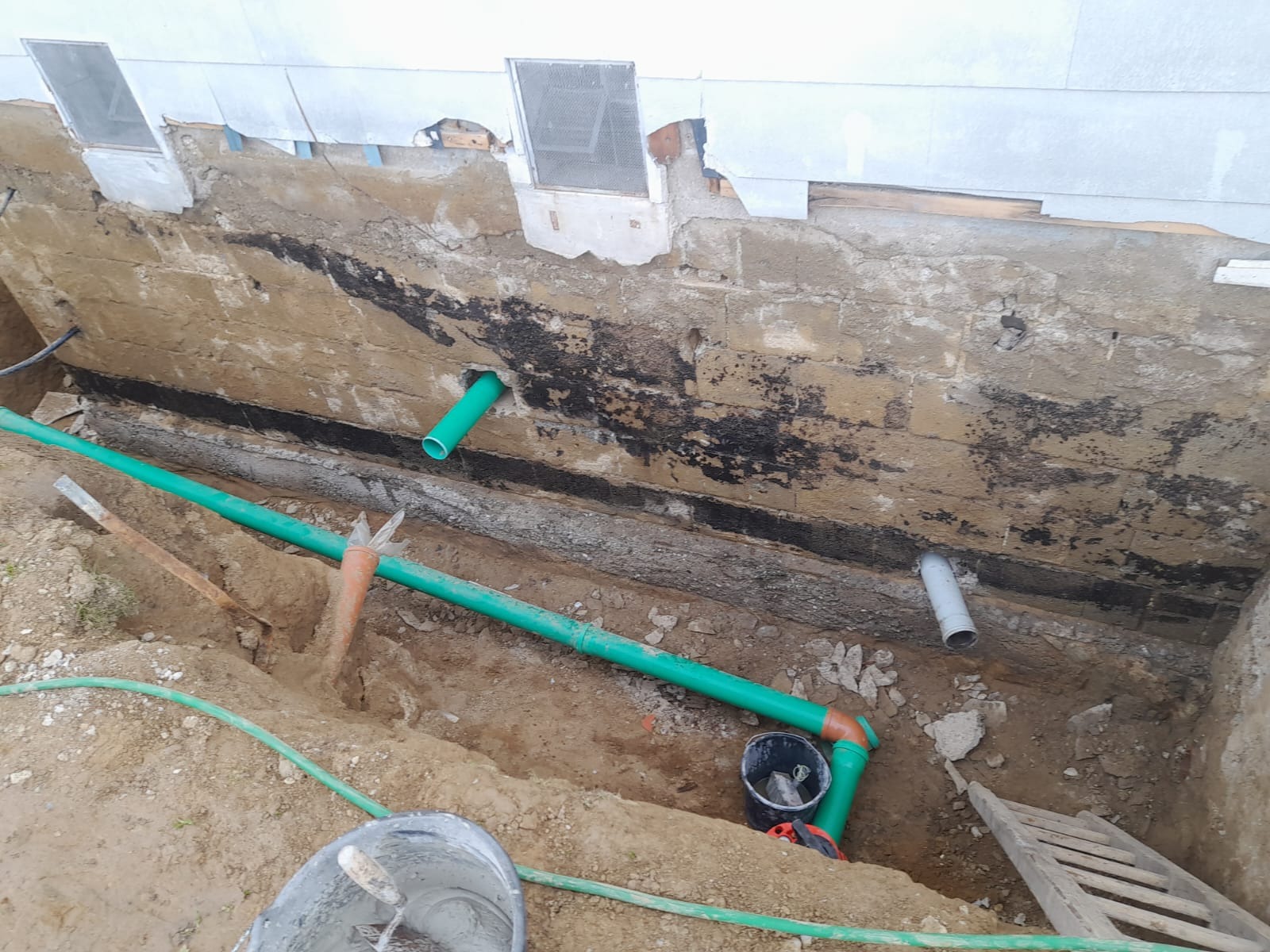 Odkopany fundament budynku z widocznymi rurami kanalizacyjnymi w kolorze zielonym i szarym, w wykopie leżą narzędzia i wiadro z zaprawą murarską, ściana fundamentu z widocznymi śladami wilgoci...