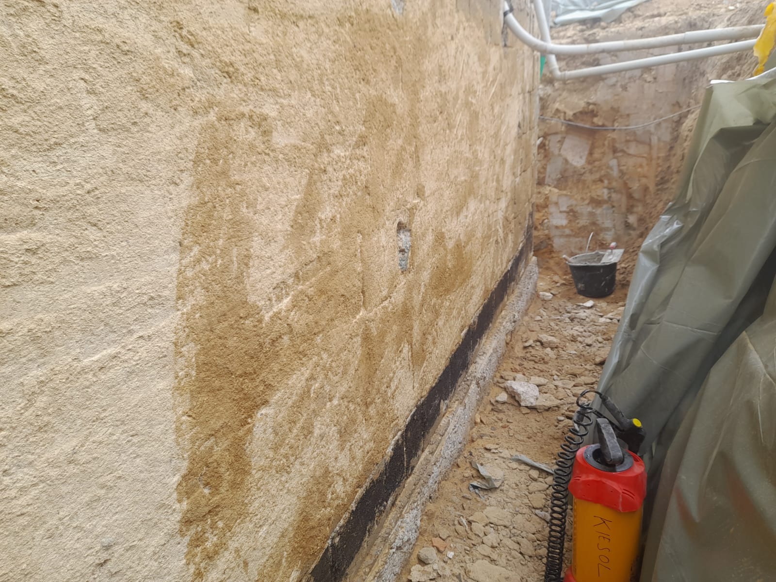 Świeżo zaizolowana ściana fundamentowa z widocznymi śladami wilgoci i czarną hydroizolacją, w wykopie z narzędziami i wiaderkiem.