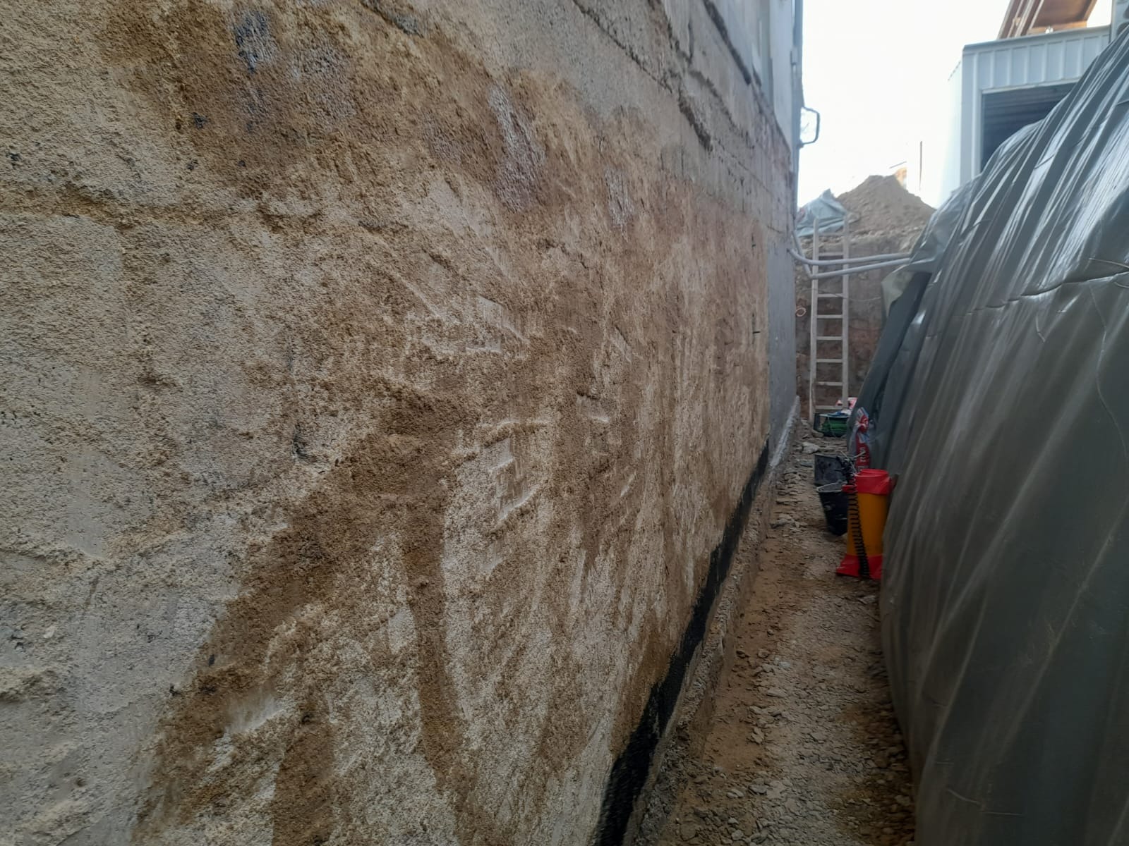 Ściana fundamentowa z widoczną izolacją przeciwwilgociową i śladami ziemi, wykop wzdłuż ściany, drabina oparta o wykop w tle.