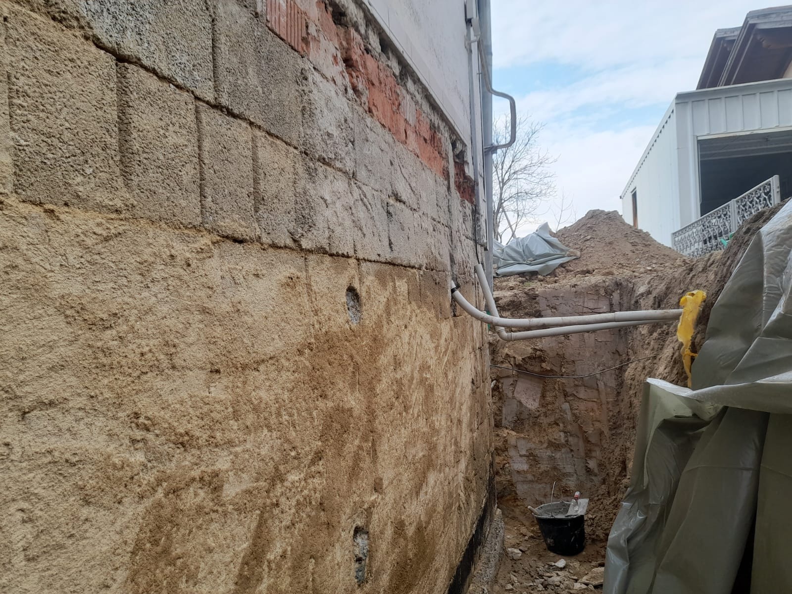 Odkopany fundament budynku z widocznymi śladami wilgoci, fragmentem odsłoniętej cegły oraz rurami instalacji. W wykopie widoczne wiadro i pryzma ziemi.