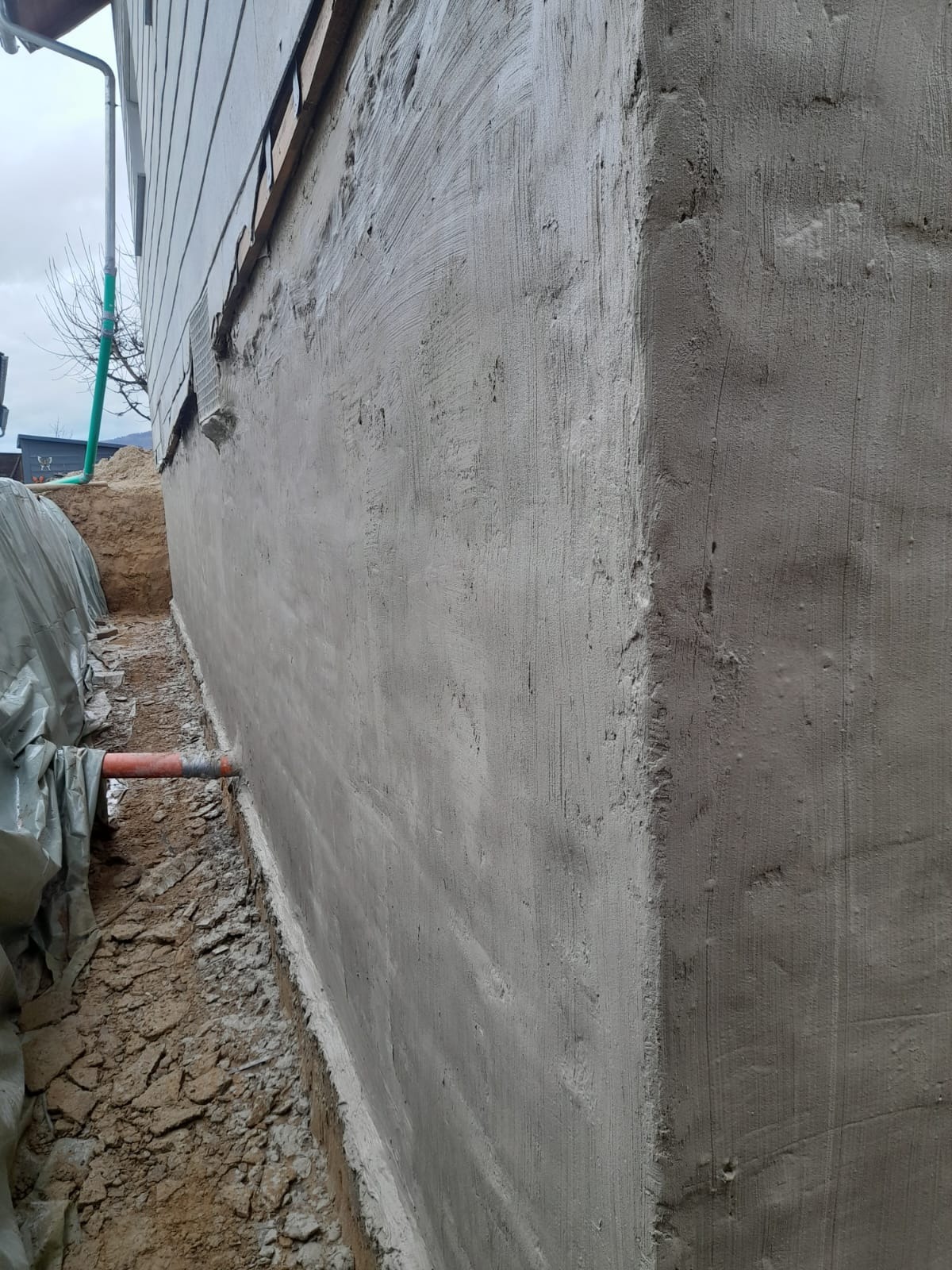 Świeżo otynkowana ściana fundamentowa budynku z widoczną warstwą izolacji przeciwwilgociowej na styku z gruntem, częściowo zakryta folią ochronną. Widoczny fragment rury odprowadzającej wodę.