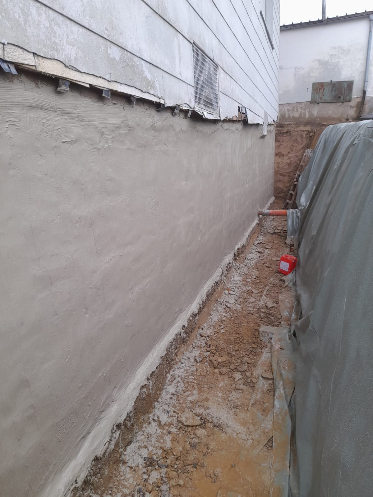 Świeżo otynkowana ściana fundamentowa z widocznym poziomem gruntu obok budynku, zabezpieczona folią i z czerwoną puszką farby w pobliżu.