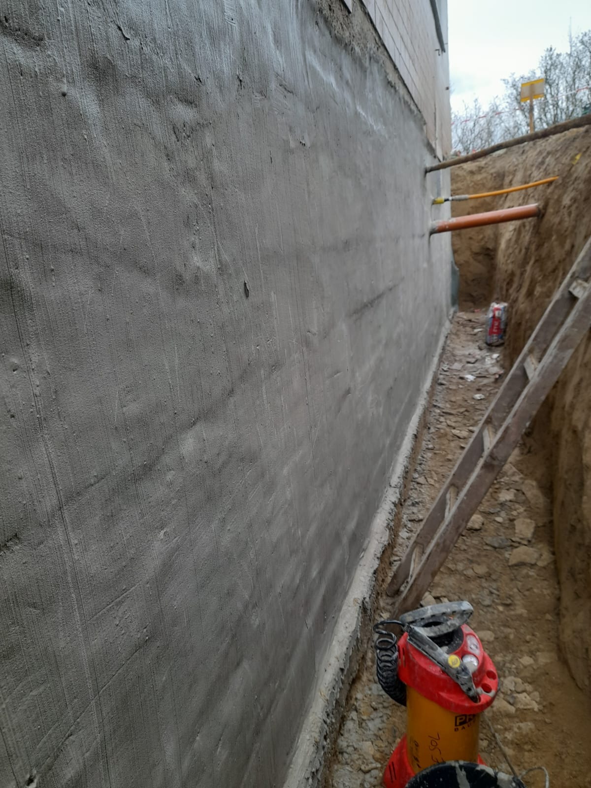 Świeżo zaizolowana, szara ściana fundamentowa budynku, widoczna z wykopu. W tle rury przechodzące przez ścianę. Obok drabina oparta o ścianę wykopu i narzędzia budowlane.