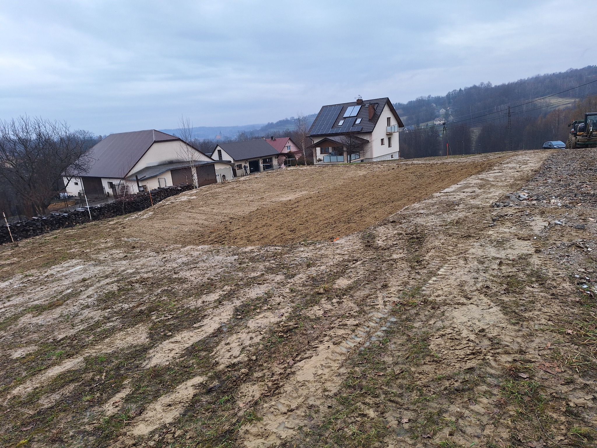 Przygotowany teren pod budowę z widocznymi śladami opon, otoczony domami w krajobrazie pagórkowatym, pochmurne niebo.