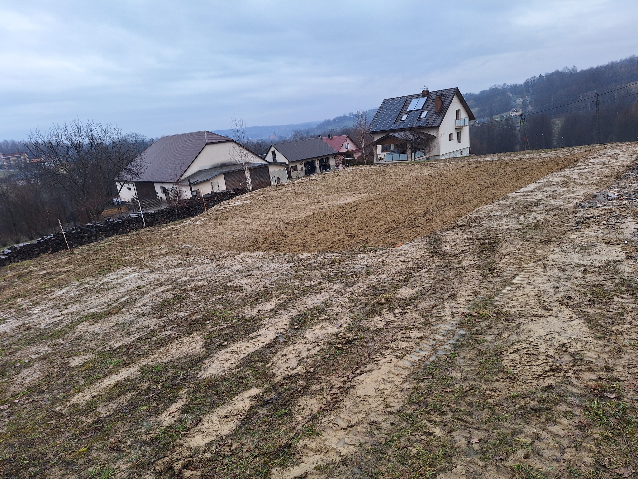 Przygotowany teren pod budowę z widocznymi śladami opon, w tle zabudowa jednorodzinna i zalesione wzgórza w pochmurny dzień.