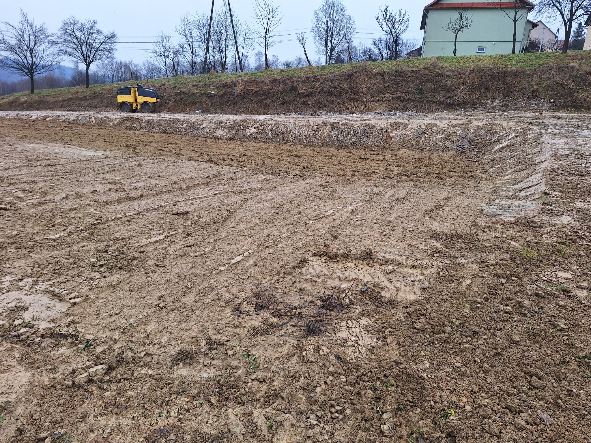 Teren przygotowany pod budowę z widocznymi śladami opon i ubijarką w tle, na wzgórzu widać budynek mieszkalny.
