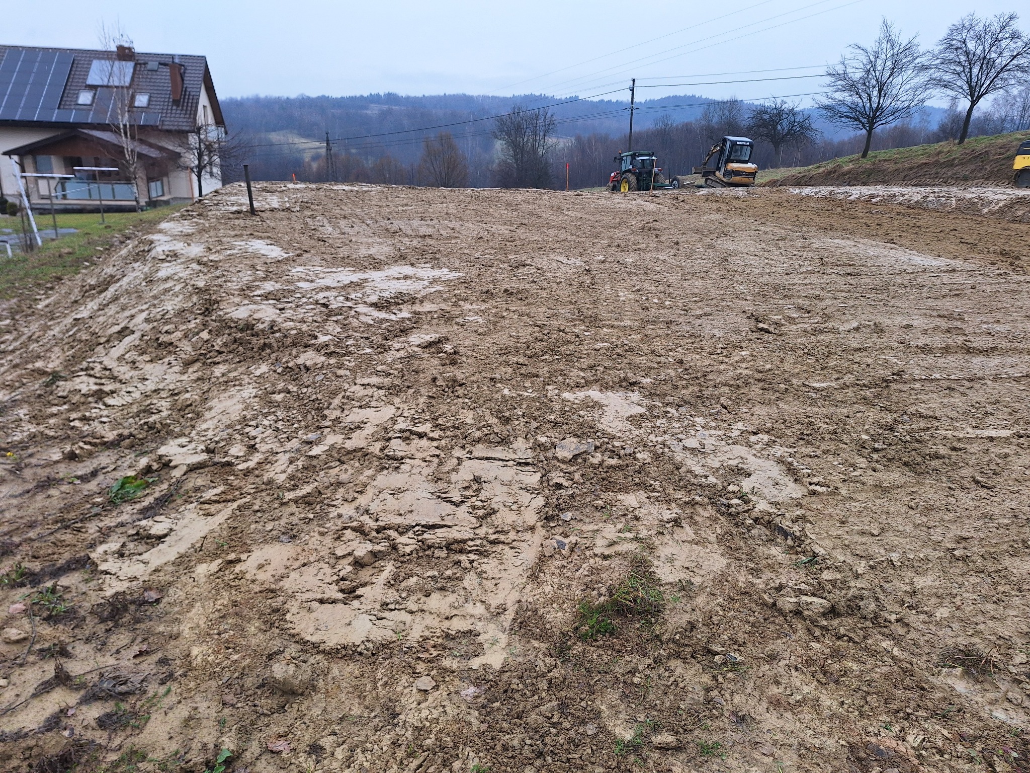 Teren budowy z ciężkim sprzętem, w tle dom z panelami słonecznymi i zalesione wzgórza, ziemia świeżo wyrównana, błotnista.
