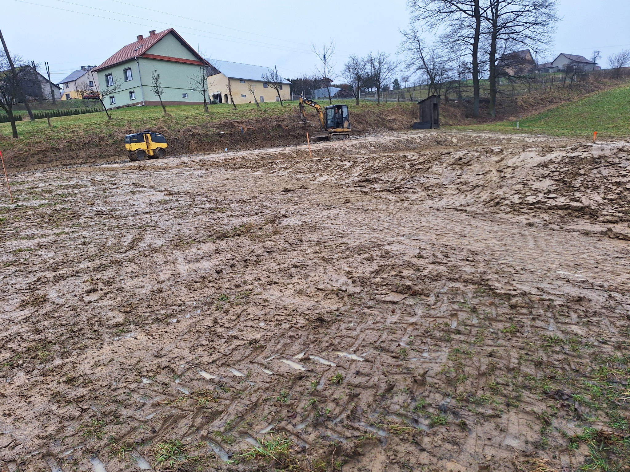 Przygotowanie terenu pod budowę: walec i koparka pracują na rozległym, błotnistym placu w pochmurny dzień. Widoczne ślady opon i gąsienic w grząskim gruncie.