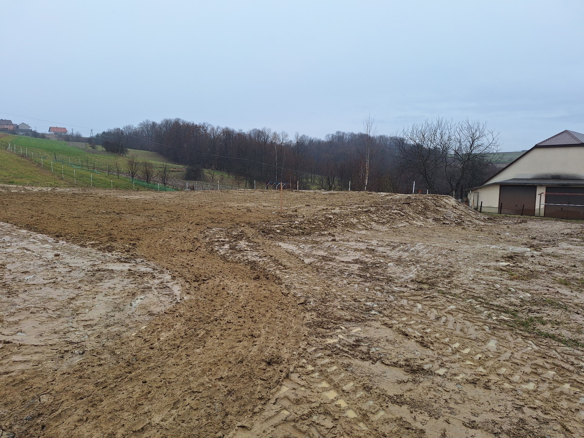 Rozjeżdżony, błotnisty teren po robotach ziemnych, widoczne ślady opon, w tle budynek gospodarczy i zalesione wzgórze pod pochmurnym niebem.