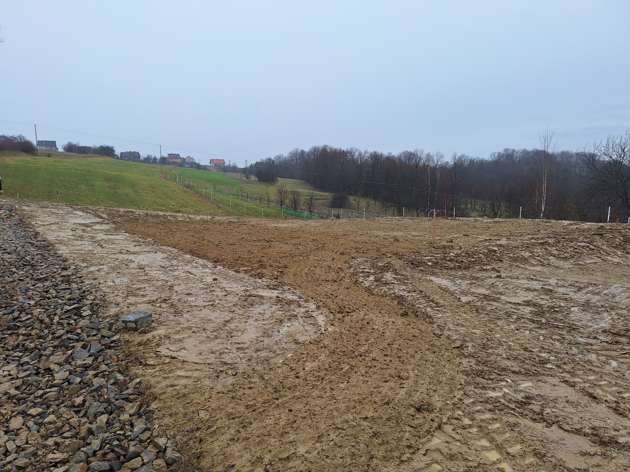 Przygotowany teren pod budowę na obszarze wiejskim, widoczne ślady opon i kamienie na obrzeżach, w tle domy na wzgórzu i pochmurne niebo.