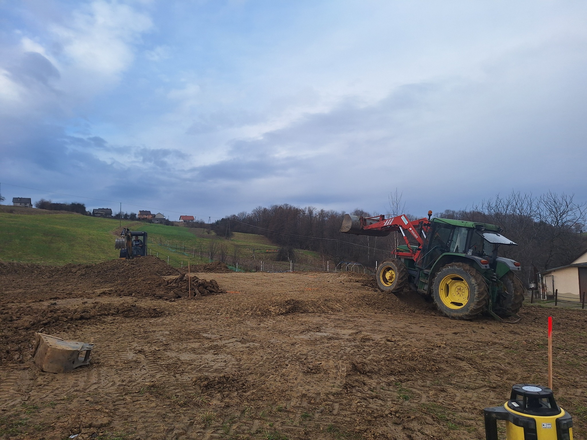 Przygotowanie terenu pod budowę: koparka i traktor z łyżką na błotnistym placu budowy, z widocznym laserowym niwelatorem i odłożoną łyżką koparki w tle.