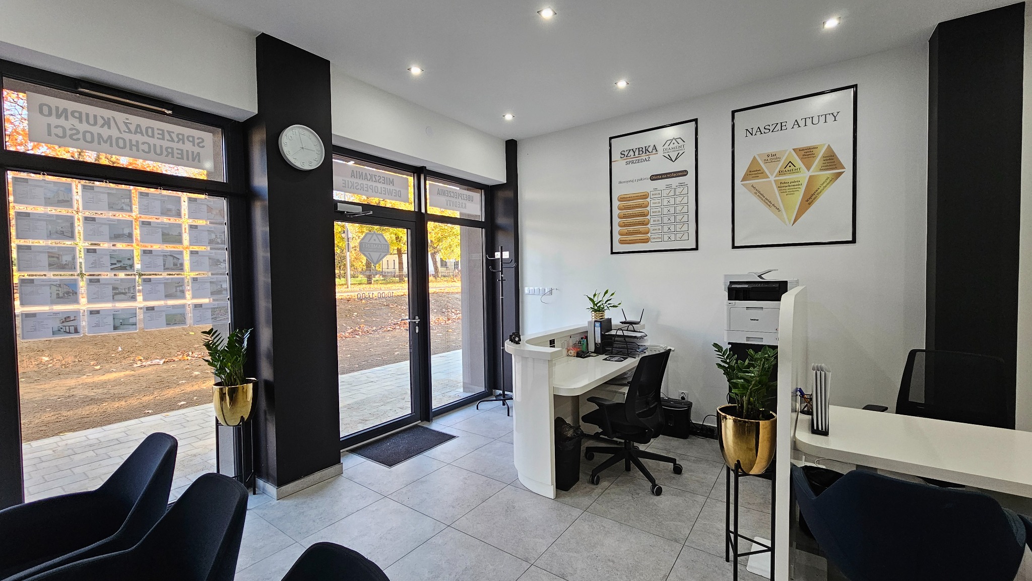 Wnętrze biura agencji nieruchomości z recepcją, plakatami informacyjnymi na ścianach i ofertami w oknie wystawowym. Widoczne krzesła dla klientów i rośliny w złotych donicach.