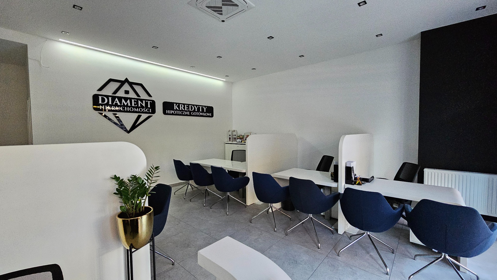 Nowoczesne biuro nieruchomości z logo 'Diament Nieruchomości' i 'Kredyty Hipoteczne Gotówkowe' na ścianie, białe biurka, niebieskie krzesła, roślina w złotej doniczce przy recepcji.
