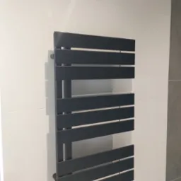 Czarny, poziomy grzejnik drabinkowy na białej ścianie z płytkami. Nowoczesny design, fragment szarej ściany obok. Oświetlenie górne.