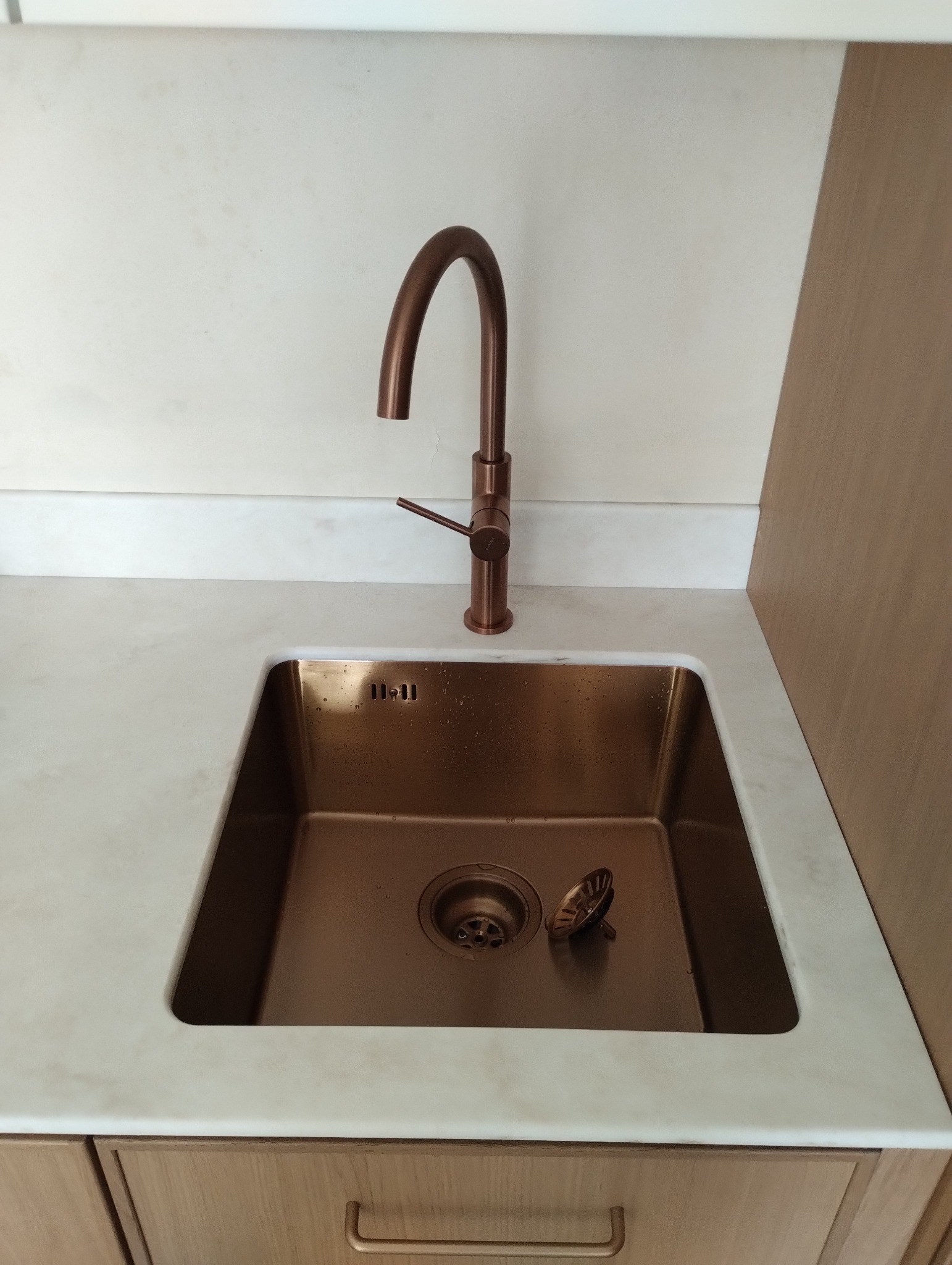 Miedziana bateria i zlew w nowej kuchni. Armatura w kolorze miedzi na blacie z jasnego marmuru, widok na odpływ i fragment szafki pod zlewem.