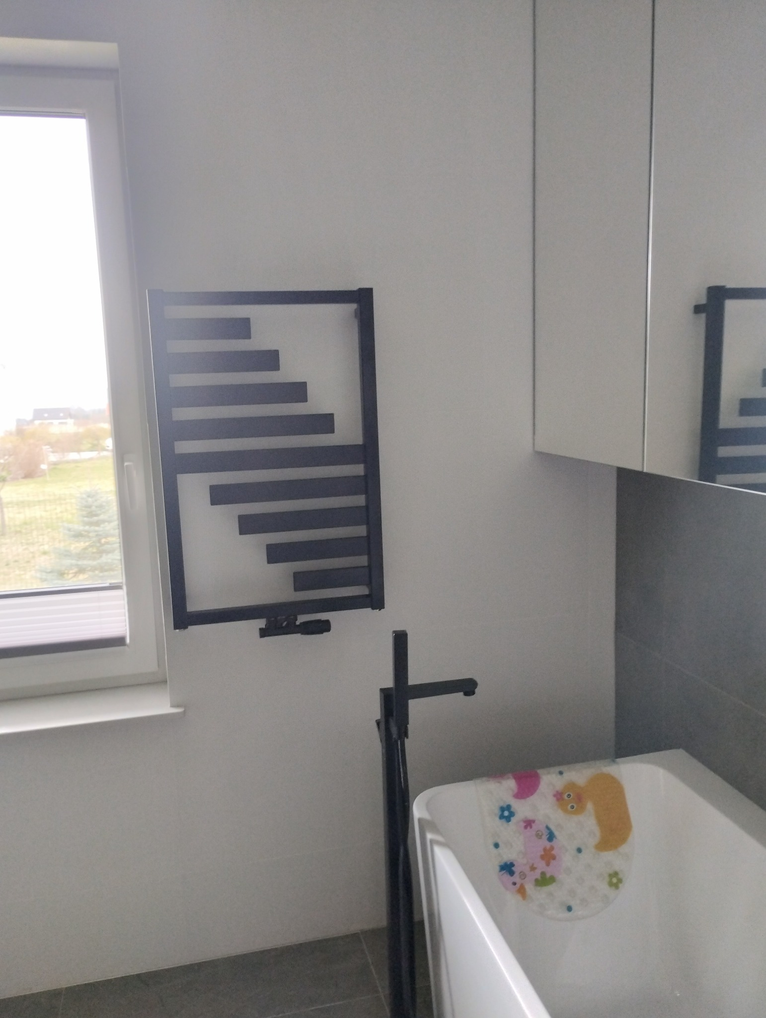 Nowoczesny, czarny grzejnik łazienkowy drabinkowy, zamontowany na białej ścianie obok okna. Widoczna wanna z antypoślizgową nakładką i bateria. Minimalistyczny design.