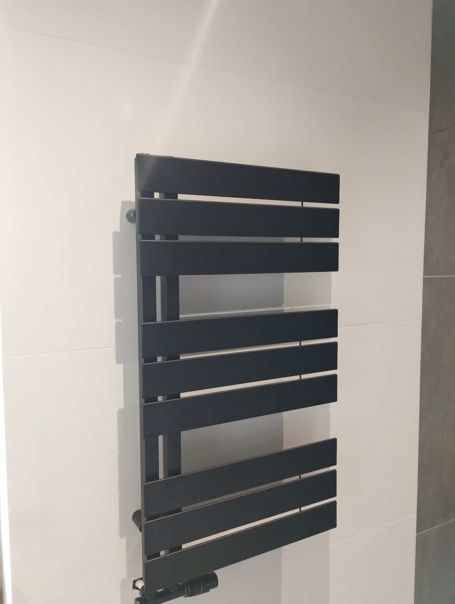 Czarny, poziomy grzejnik drabinkowy na białej ścianie z płytkami. Nowoczesny design, fragment szarej ściany obok. Oświetlenie górne.