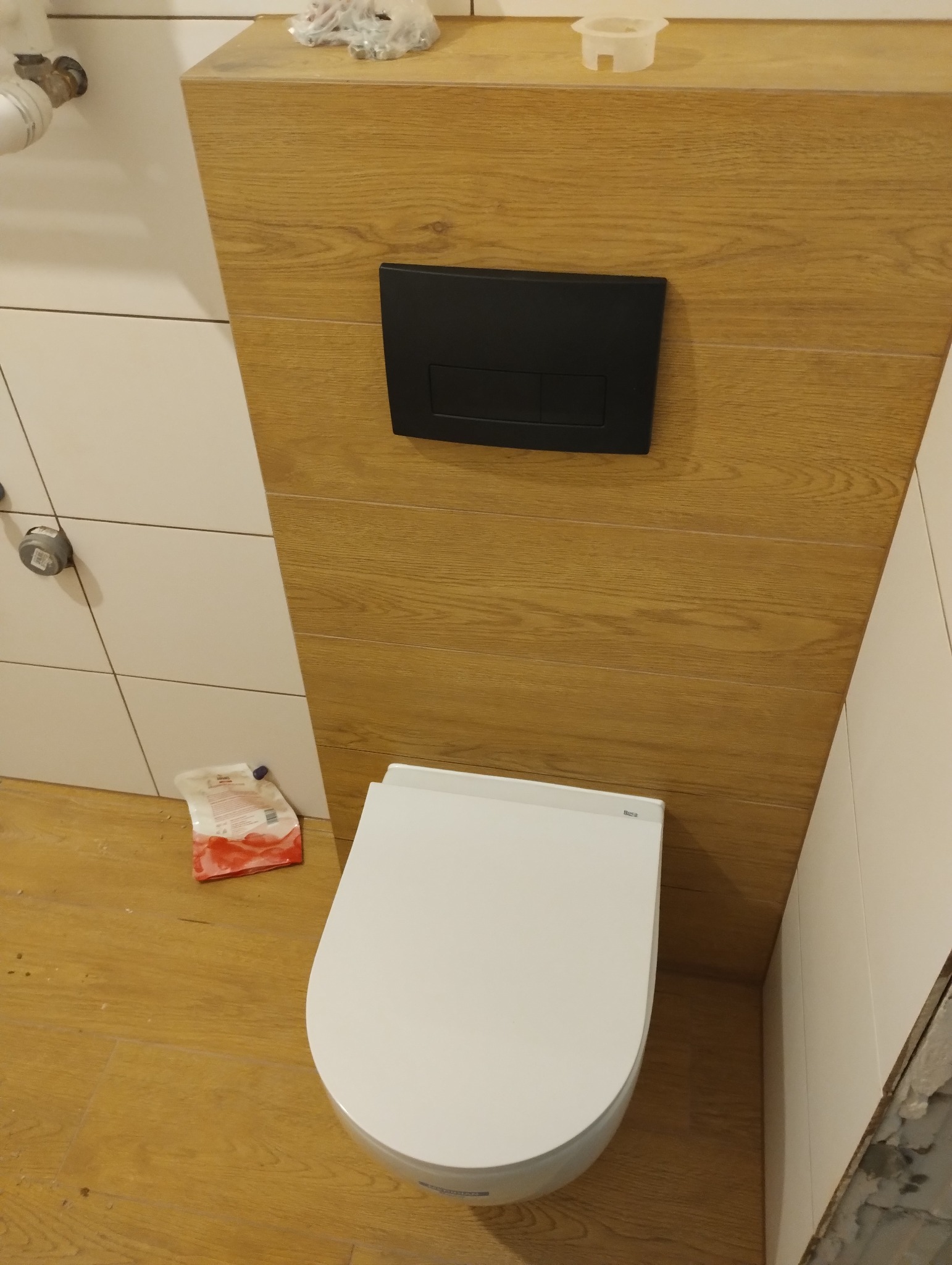 Nowoczesna toaleta podwieszana z czarnym przyciskiem spłukującym na tle drewnopodobnej obudowy, w trakcie montażu. Widoczne akcesoria instalacyjne.