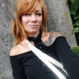 Kobieta z rudymi włosami i tatuażem na ramieniu, w czarnej bluzce z odkrytymi ramionami, pozuje na tle betonowej ściany i zielonych liści.