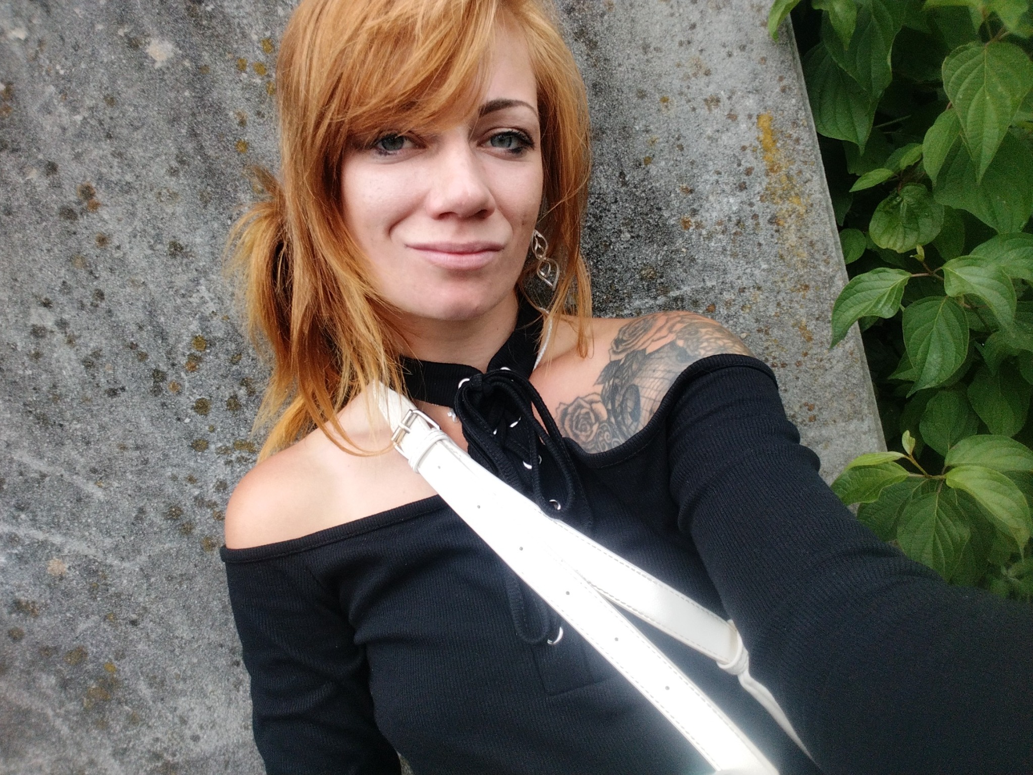 Kobieta z rudymi włosami i tatuażem na ramieniu, w czarnej bluzce z odkrytymi ramionami, pozuje na tle betonowej ściany i zielonych liści.