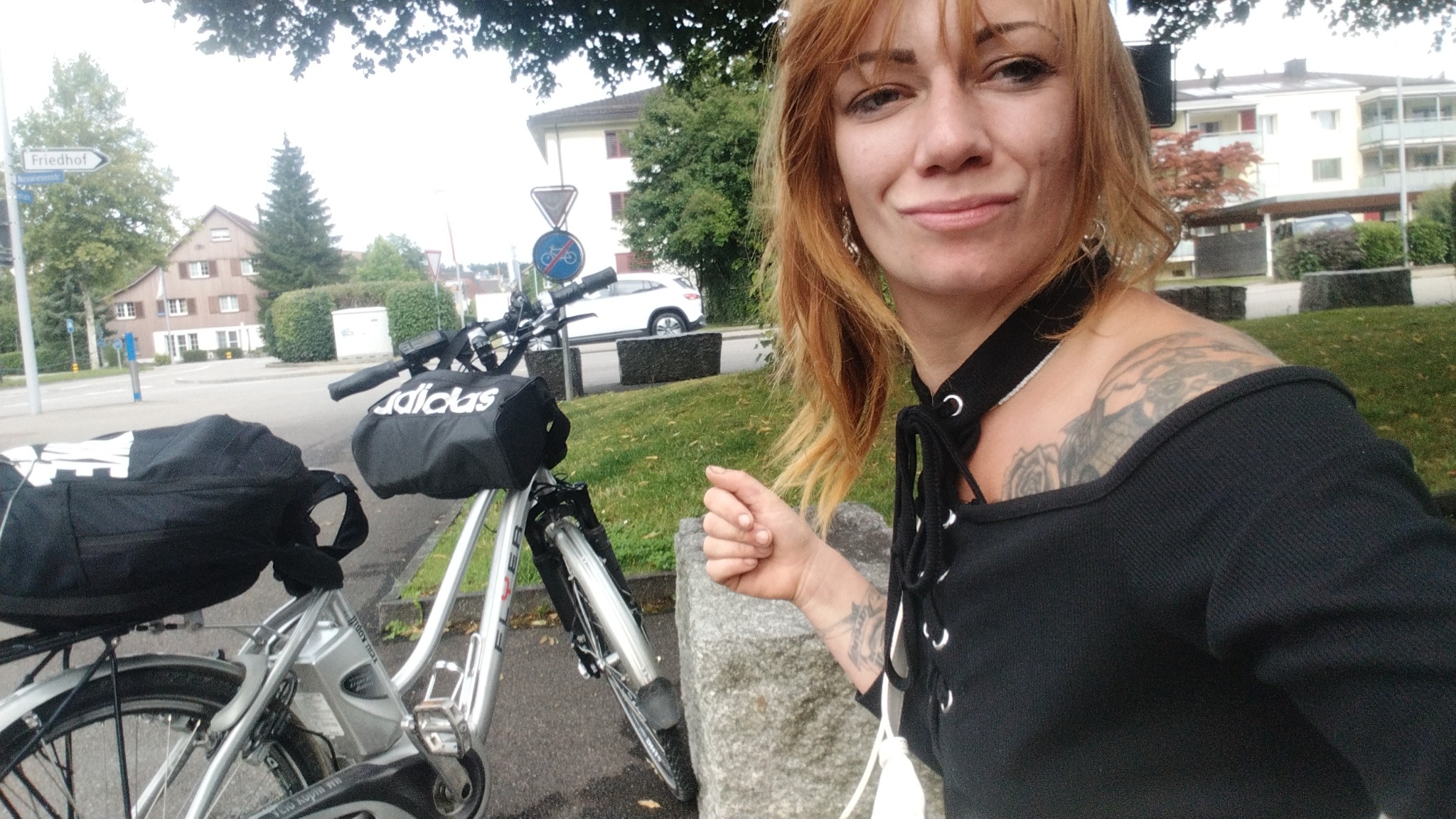 Kobieta z rowerem elektrycznym Flyer, obok torba Adidas. W tle budynki mieszkalne i znak drogowy. Uśmiechnięta kobieta z tatuażami na ramieniu.