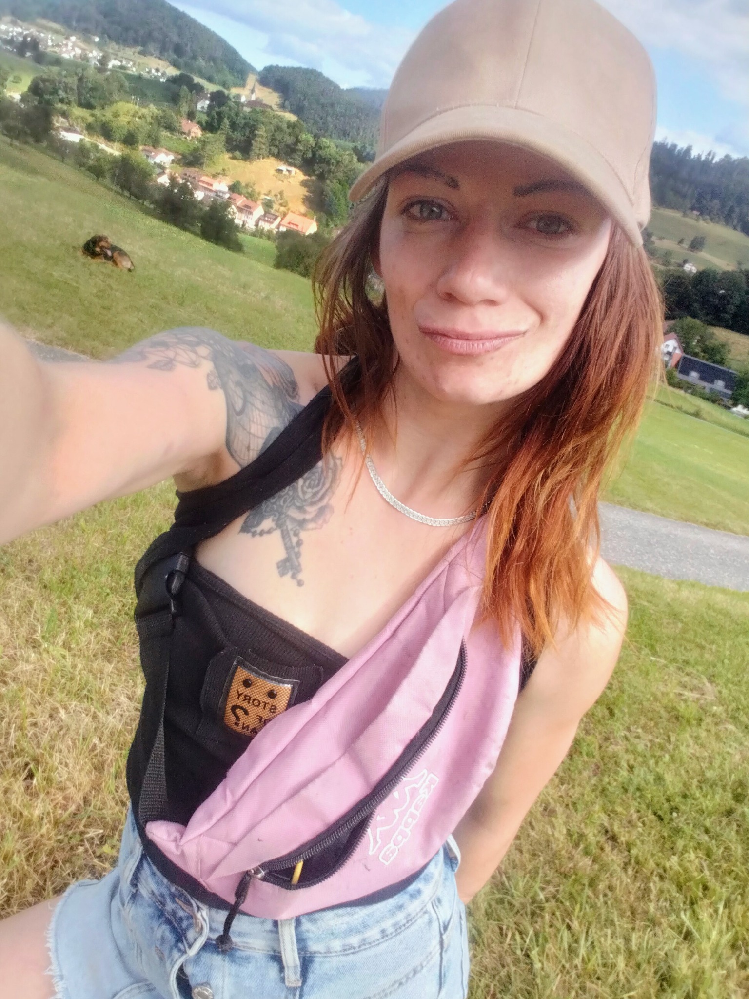 Kobieta w czapce i różowej nerce na tle zielonego wzgórza z zabudowaniami w Legnicy. Widoczny tatuaż na ramieniu, trawa i ścieżka.