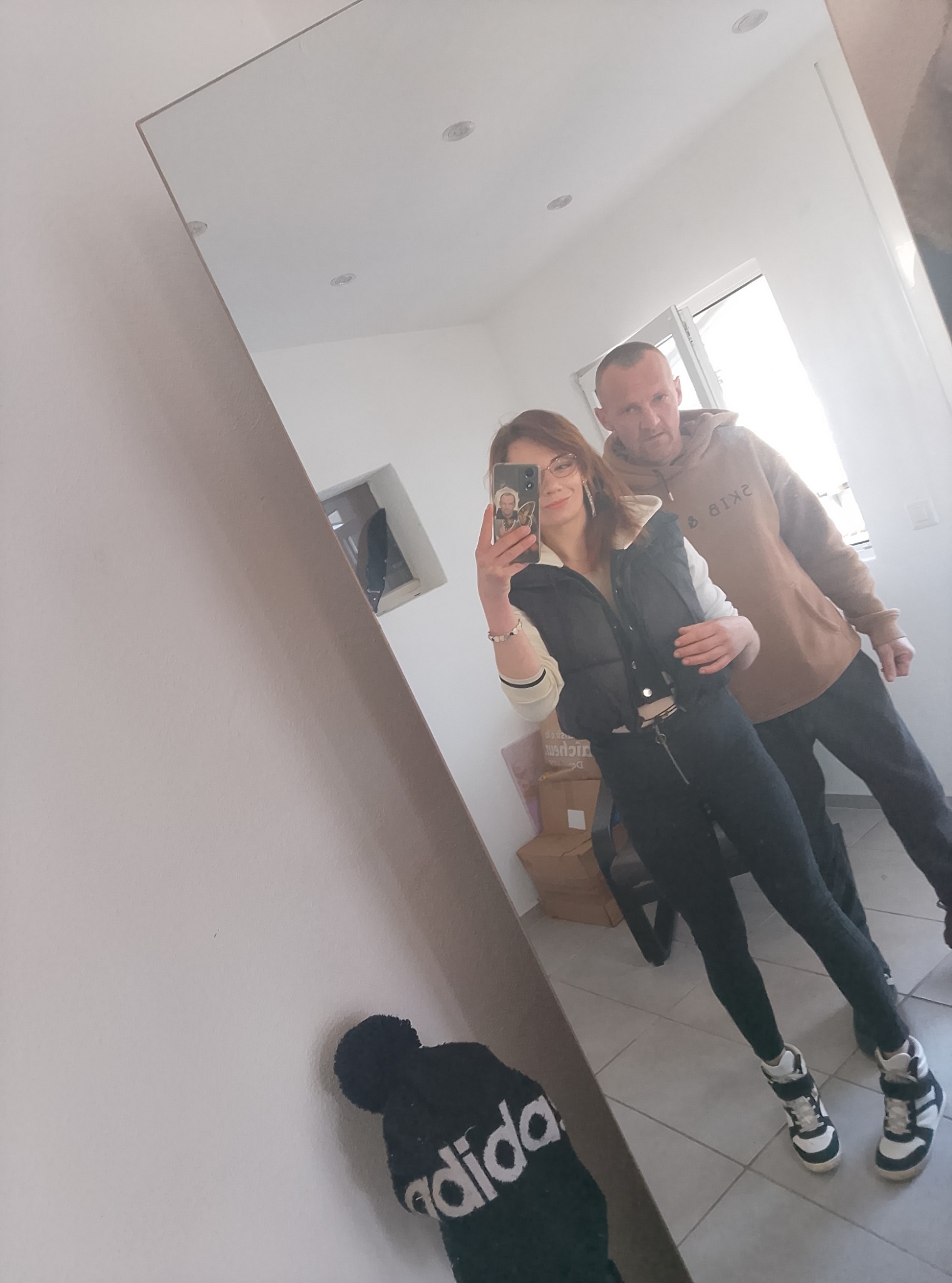 Para pozuje do selfie w lustrze w jasnym wnętrzu. Widoczna odzież sportowa i codzienna. Ujęcie z dołu, w tle okno i kartony. Nieco zamazane odbicie.