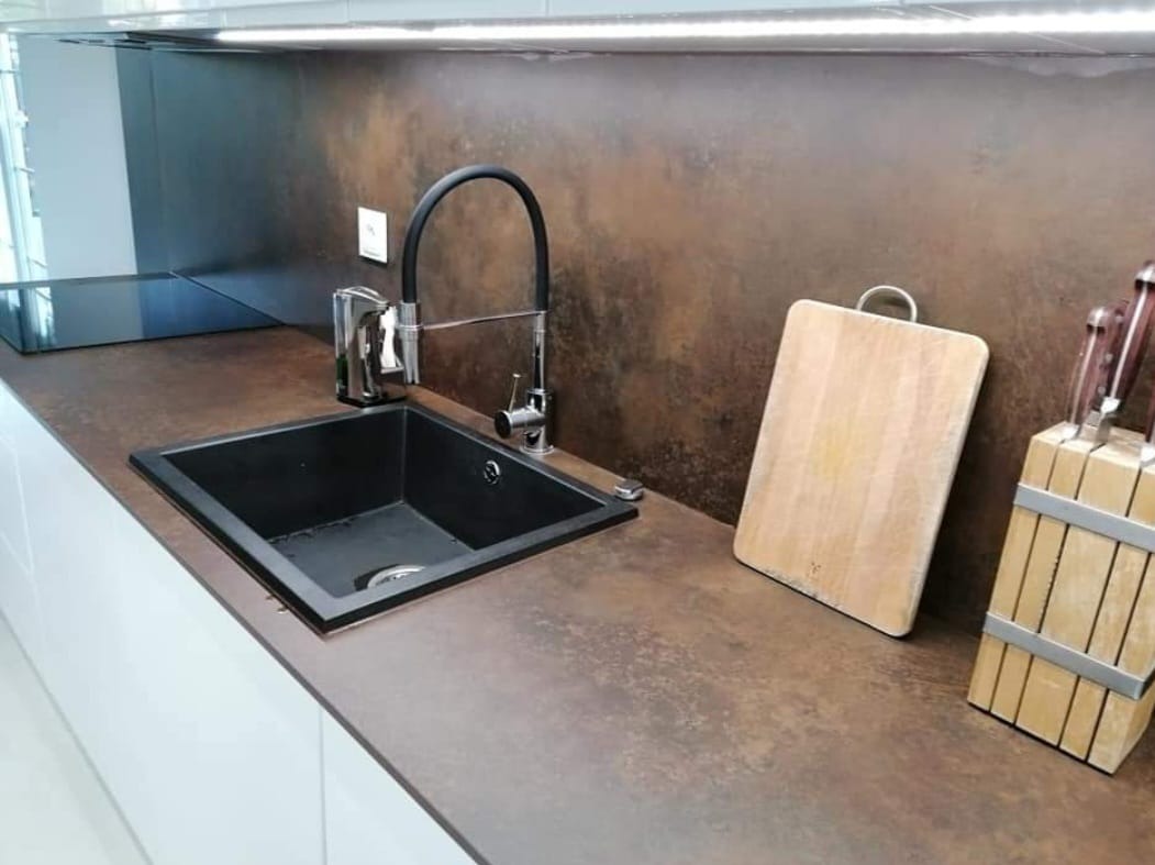 Czarny zlew wpuszczany w blat kamienny o fakturze rdzy, bateria z czarnym wężem, drewniana deska do krojenia i blok z nożami.