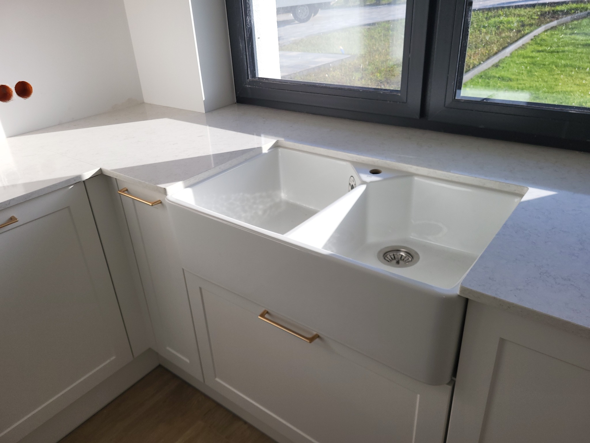 Biały, dwukomorowy zlew ceramiczny typu farmhouse wmontowany w szary blat z delikatnym wzorem, złote uchwyty szafek, widok na zewnątrz przez okno.