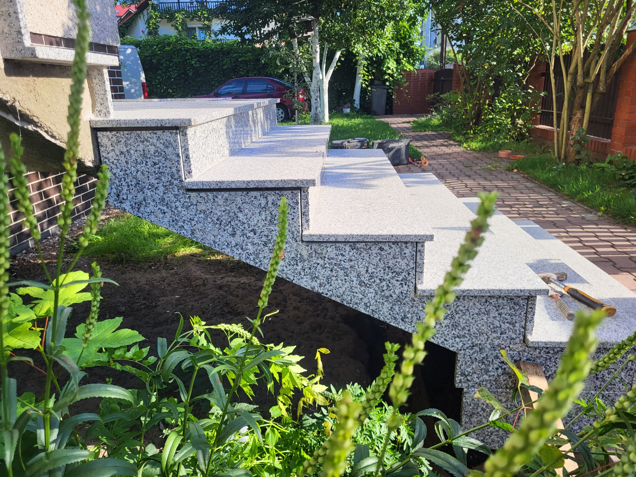 Zewnętrzne schody z szarego granitu prowadzące do domu, widoczne narzędzia montażowe, fragment ogrodu z roślinami na pierwszym planie.