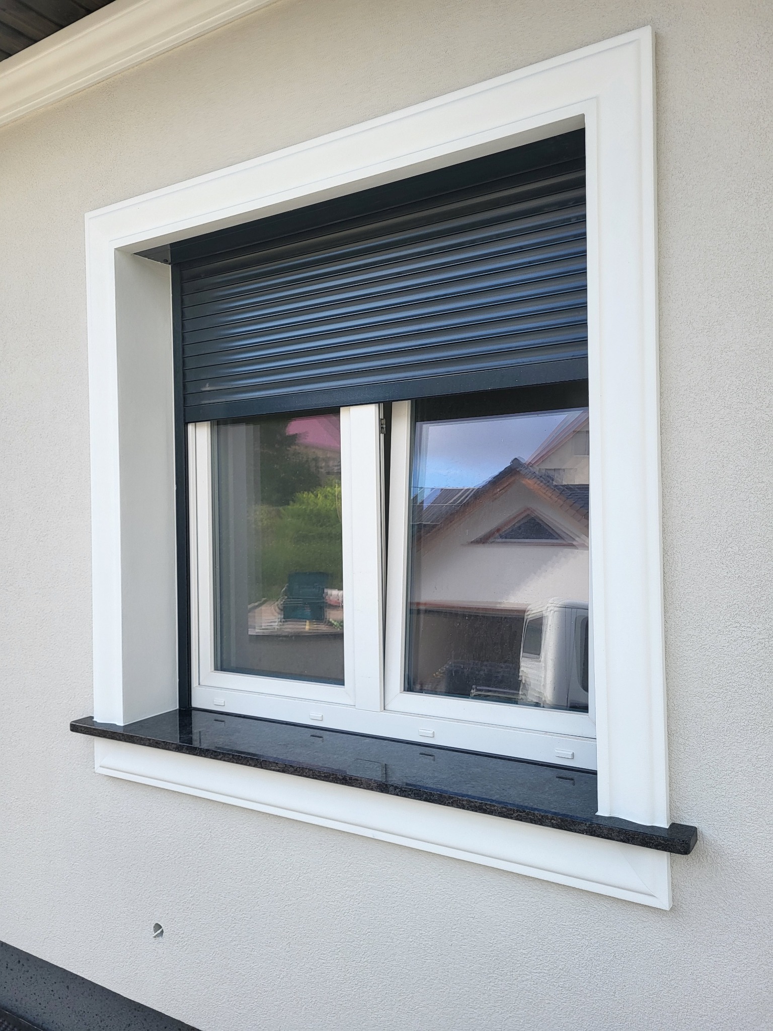 Okno z czarnym parapetem, białymi ramami i zewnętrzną roletą, widok z zewnątrz na elewację budynku.