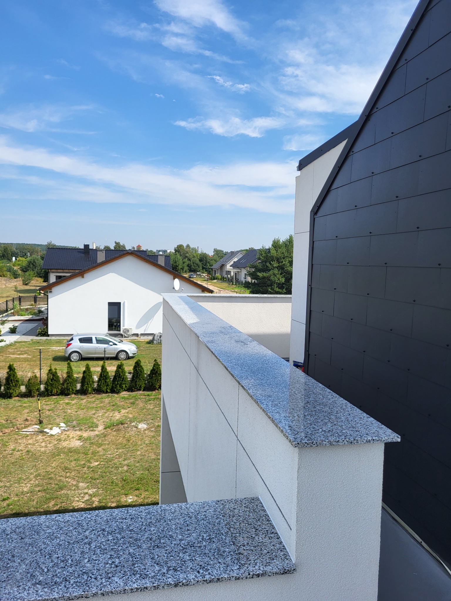 Barierka tarasu z szarego kamienia na tle nowoczesnego domu z czarną fasadą i widokiem na ogród z samochodem i tujami