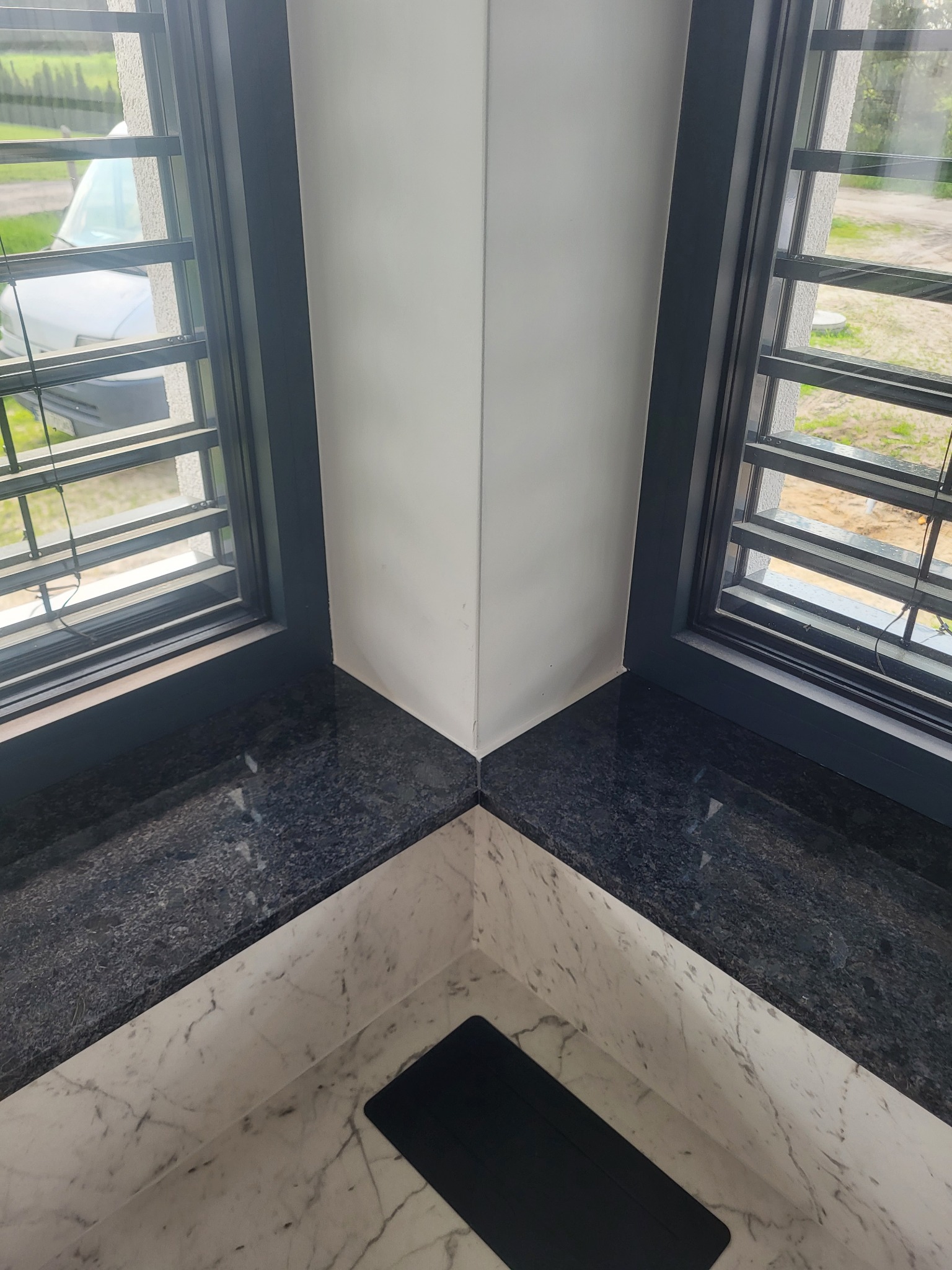 Wnętrze z dwoma oknami z ciemnymi ramami i parapetem z ciemnego granitu, kontrastującym z marmurowym wykończeniem ściany pod parapetem; widok z okna na zieleń w Zaborze.